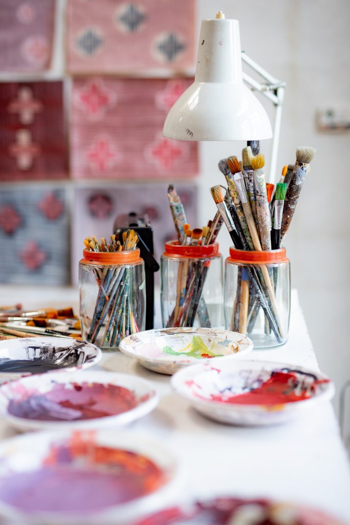 A propos : Table blanche sur laquelle sont posés des pots de pinceaux usés et des assiettes de peinture. Mur rose et blanc à l'arrière plan.